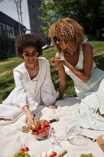Joyful african american girlfriends eating fresh strawberry and grapes during summer picnic — Stock Photo