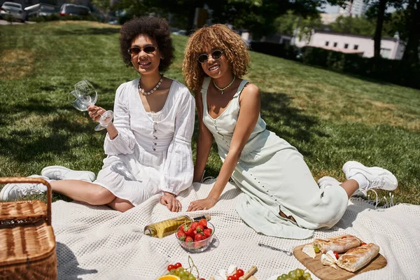African american woman holding wine glasses near girlfriend and fresh fruits, summer picnic — Stock Photo