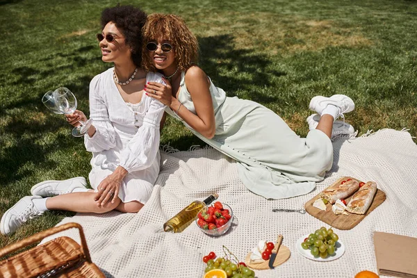 Африканская американка с бокалами вина рядом с другом и свежими фруктами на одеяле, летний пикник — стоковое фото