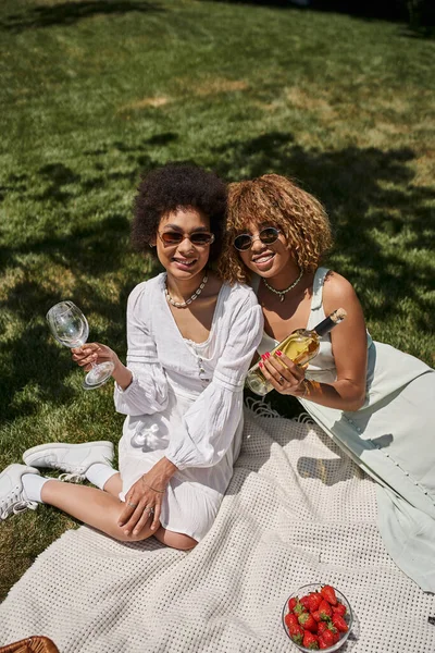 Joyful african american girlfriends, white dresses, wine bottle, glasses, summer picnic — Stock Photo