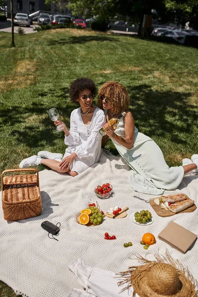 Stylish african american girlfriends on summer picnic on blanket, fresh fruits, wine, straw basket — Stock Photo