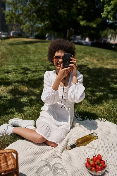 Donna afro-americana spensierata che scatta foto sulla fotocamera vintage vicino al vino e alle fragole al picnic — Foto stock