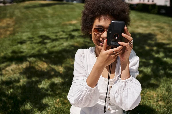 Щаслива афроамериканка в сонцезахисних окулярах фотографується на старовинній камері в літньому парку, радість — стокове фото