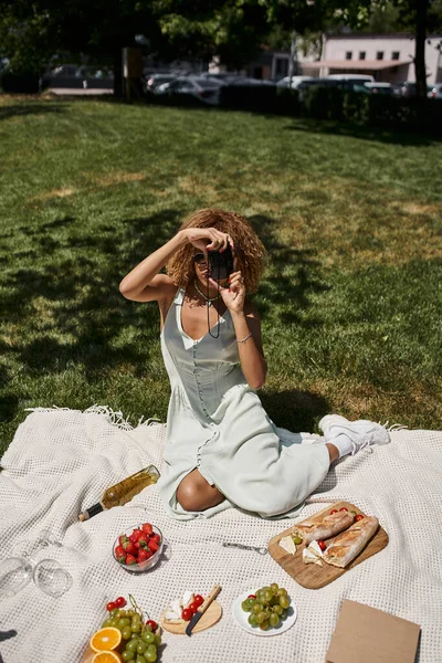 African american woman taking photo on vintage camera near wine and strawberries, summer picnic — Stock Photo