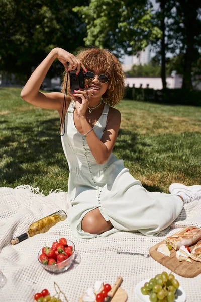 Alegre afroamericana mujer con vintage cámara tomando foto cerca de vino y comida en el picnic - foto de stock