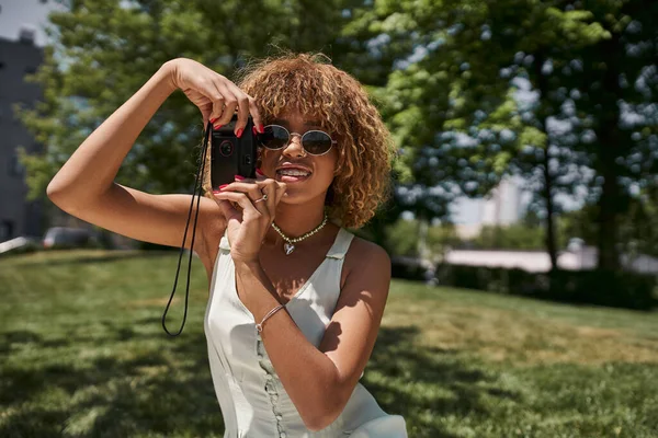 Stylish african american woman in sunglasses taking photo on vintage camera in summer park — Stock Photo