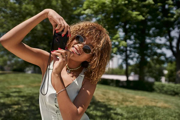 Femme afro-américaine insouciante en lunettes de soleil prendre des photos sur caméra vintage pendant les loisirs dans le parc — Photo de stock