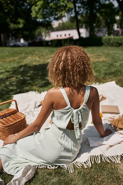 Visão traseira da jovem afro-americana sentada em cobertor perto da cesta de palha no parque — Fotografia de Stock