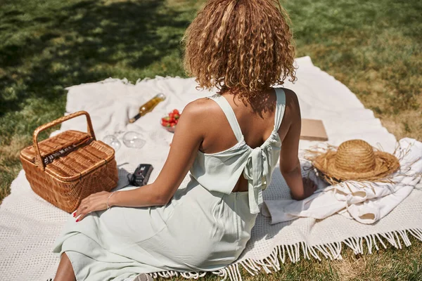 Picnic de verano en el parque, joven mujer afroamericana sentada en una manta cerca de la canasta de paja - foto de stock