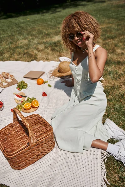 Picnic de verano, mujer afroamericana feliz sentada cerca de frutas, verduras y canasta de paja - foto de stock