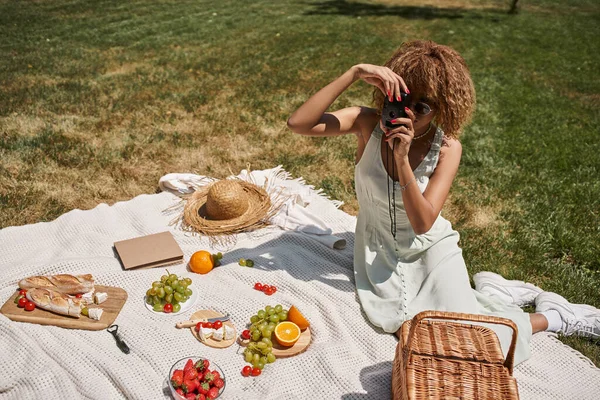 African american woman taking photo on vintage camera near fruits and vegetables, summer picnic — Stock Photo