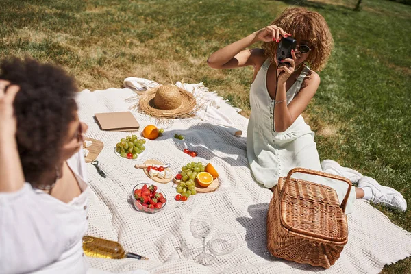 Young african american woman taking photo of girlfriend on vintage camera, picnic in summer park — Stock Photo