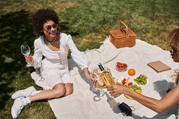 Unbekümmerte afrikanisch-amerikanische Freundinnen gießen beim Picknick Wein in die Nähe von Obst und Gemüse — Stockfoto