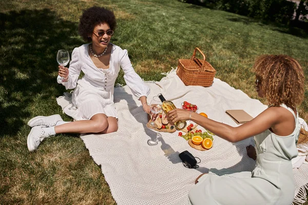 Mujer afroamericana feliz sosteniendo vasos cerca de novia vertiendo vino durante el picnic de verano - foto de stock