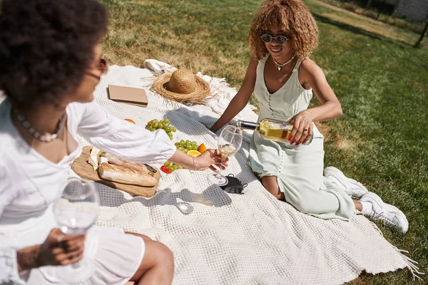 Africano americano mulher segurando copos de vinho perto feliz namorada derramando vinho — Fotografia de Stock