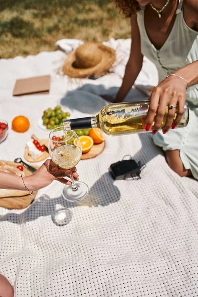 Vista cortada da mulher americana africana derramando vinho perto de frutas durante piquenique com a namorada — Fotografia de Stock