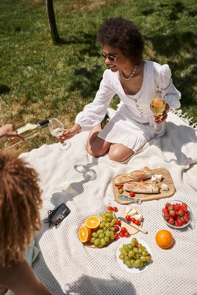 Carefree african american woman holding glass while girlfriend pouring wine on summer picnic — Stock Photo