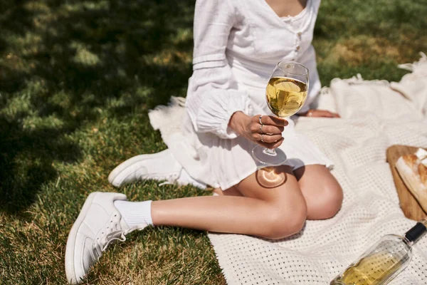 Vista recortada de la joven afroamericana sentada con una copa de vino en una manta y césped en el parque - foto de stock