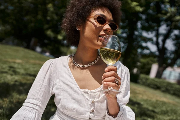 Mujer afroamericana con estilo en gafas de sol sosteniendo copa de vino en el verde parque de verano - foto de stock