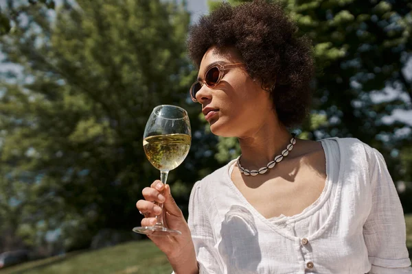 Femme afro-américaine branchée dans les lunettes de soleil boire du vin sur le pique-nique dans le parc — Photo de stock