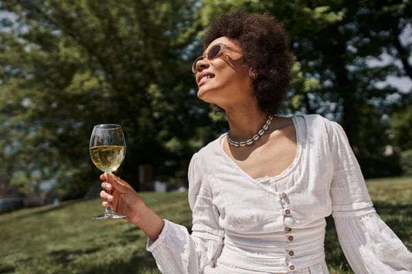 Mulher afro-americana despreocupada e elegante em óculos de sol segurando copo de vinho no parque de verão — Fotografia de Stock