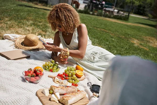 Donna afroamericana con bicchiere di vino utilizzando smartphone vicino a frutta e fidanzata sul pic-nic nel parco — Foto stock