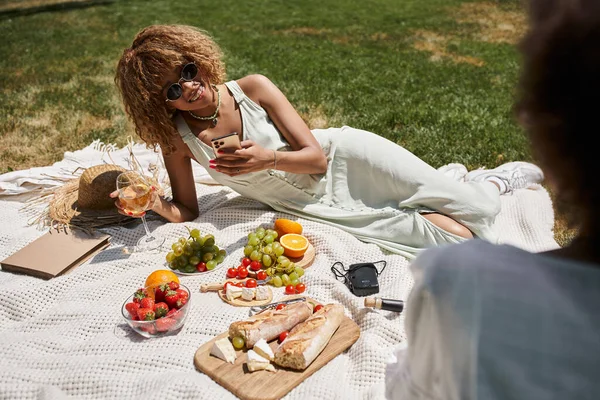 Alegre mujer afroamericana con teléfono inteligente y copa de vino cerca de novia y frutas en el parque - foto de stock