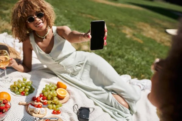 Sorridente mulher afro-americana segurando smartphone com tela em branco durante piquenique no parque de verão — Fotografia de Stock