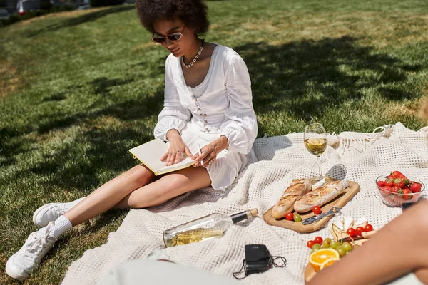Mulher afro-americana ler livro perto de vinho e comida em cobertor no parque, piquenique de verão — Fotografia de Stock