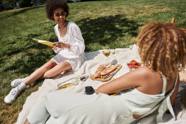 Mujer afroamericana feliz leyendo libro cerca de novia, comida y vino en el picnic en el parque de verano - foto de stock