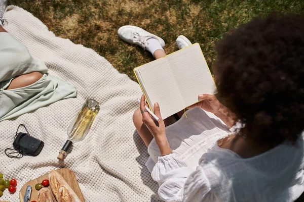 Vista superior de la mujer afroamericana con cuaderno cerca de novia y botella de vino, picnic de verano - foto de stock