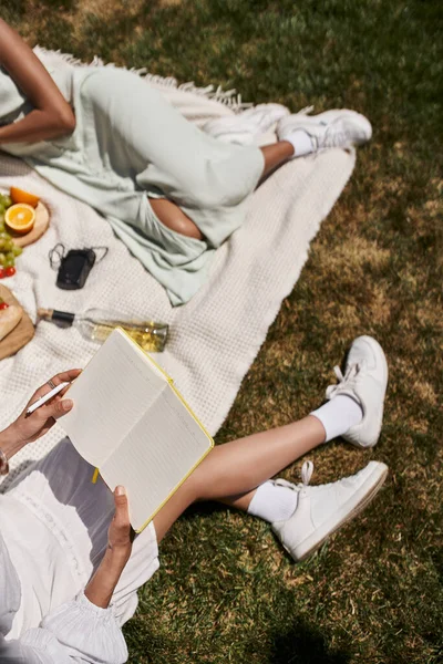 African american woman with notebook near girlfriend, fruits and wine bottle on blanket in park — Stock Photo
