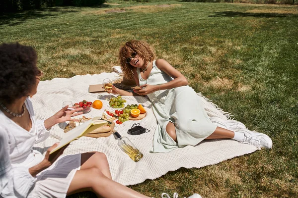 Mujer afroamericana con teléfono inteligente y copa de vino hablando con su novia en el picnic de verano - foto de stock