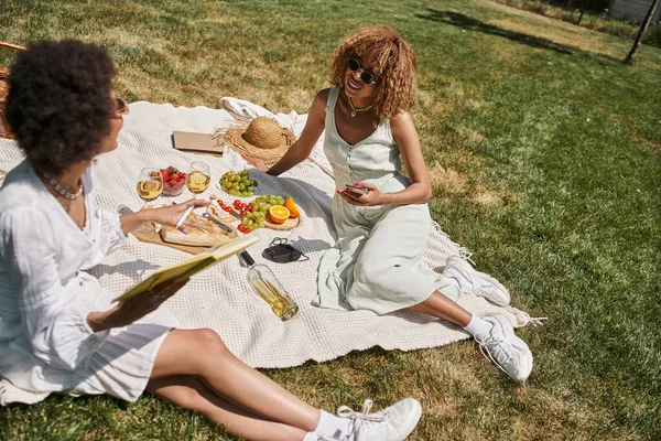 Summer picnic, african american woman with smartphone talking to girlfriend near food and wine — Stock Photo