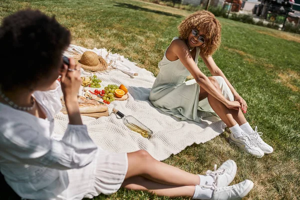 African american woman taking photo of girlfriend on vintage camera, picnic in summer park — Stock Photo