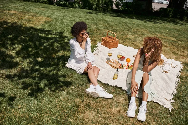 Mujer afroamericana con cámara vintage tomando fotos de su novia en el césped durante el picnic de verano - foto de stock