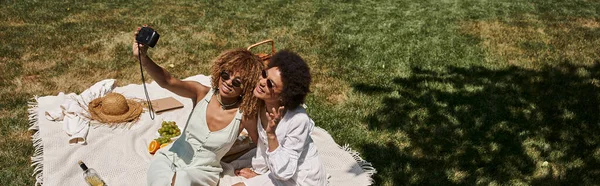 Cheerful african american girlfriends taking selfie on vintage camera on blanket near fruits in park — Stock Photo
