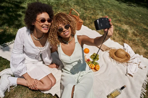 Безтурботні афроамериканські подружки в сонцезахисних окулярах беруть селфі на старовинну камеру, пікнік, банер — стокове фото