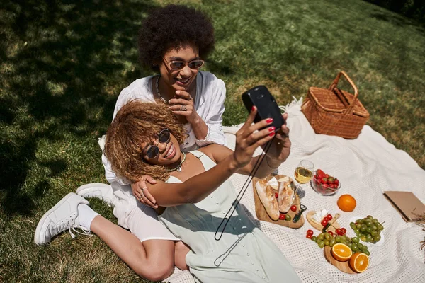 Вид сверху на веселых африканских американских подружек, делающих селфи возле еды и вина, пикник в парке — стоковое фото