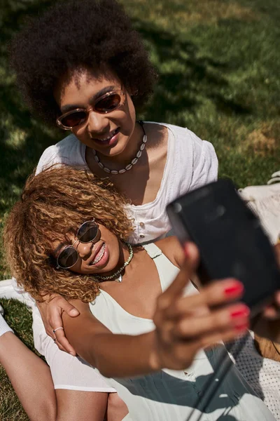 Happy and stylish african american girlfriends in sunglasses taking selfie on vintage camera in park — Stock Photo