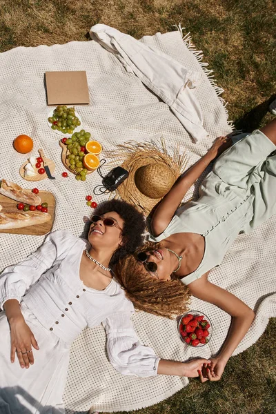 Vista superior de las novias afroamericanas positivas cogidas de la mano y colocadas sobre una manta durante el picnic - foto de stock