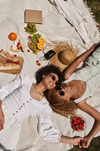 Vue du dessus des femmes afro-américaines heureuses allongées sur une couverture près du chapeau de paille et de la nourriture, pique-nique d'été — Photo de stock