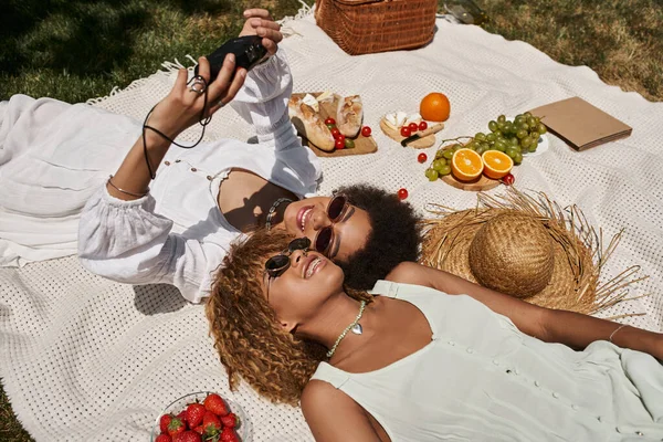 African american women taking selfie on vintage camera near food on blanket, summer picnic, joy — Stock Photo
