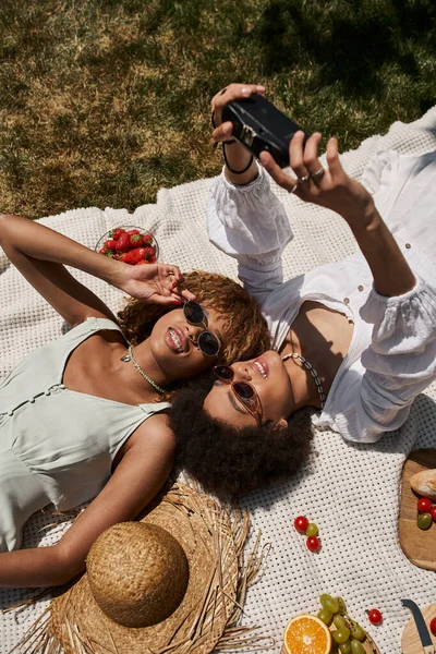 Despreocupado Africano americano namoradas tomando selfie na câmera vintage perto de morango fresco, piquenique — Fotografia de Stock