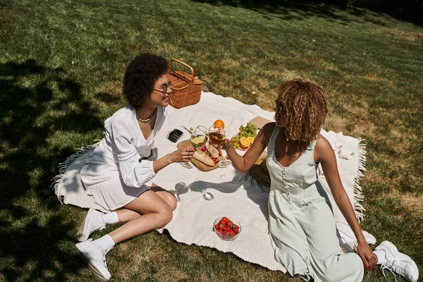 Carefree african american girlfriends clinking wine glasses near snacks on blanket, summer picnic — Stock Photo