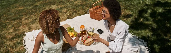 Joyful african american girlfriends clinking wine glasses near food, summer picnic in park, banner — Stock Photo