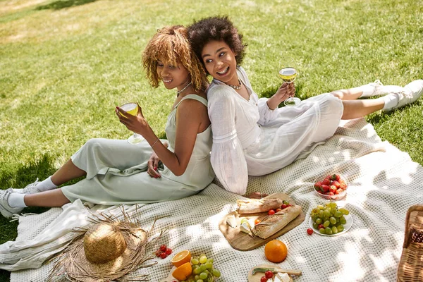 Novias afroamericanas con copas de vino sentadas espalda con espalda cerca de la comida en el picnic de verano - foto de stock