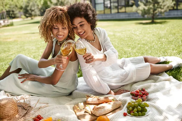 Novias afroamericanas sentadas espalda con espalda, tintineo copas de vino cerca de aperitivos, picnic de verano - foto de stock