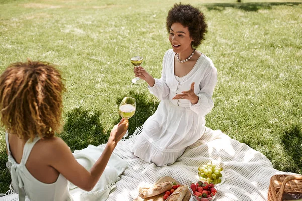 Mulher americana africana animado com copo de vinho falando com a namorada, comida em cobertor, piquenique de verão — Fotografia de Stock