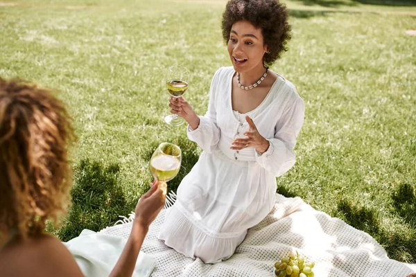 Femme américaine africaine émotionnelle avec verre de vin parlant à la petite amie pendant le pique-nique d'été dans le parc — Photo de stock